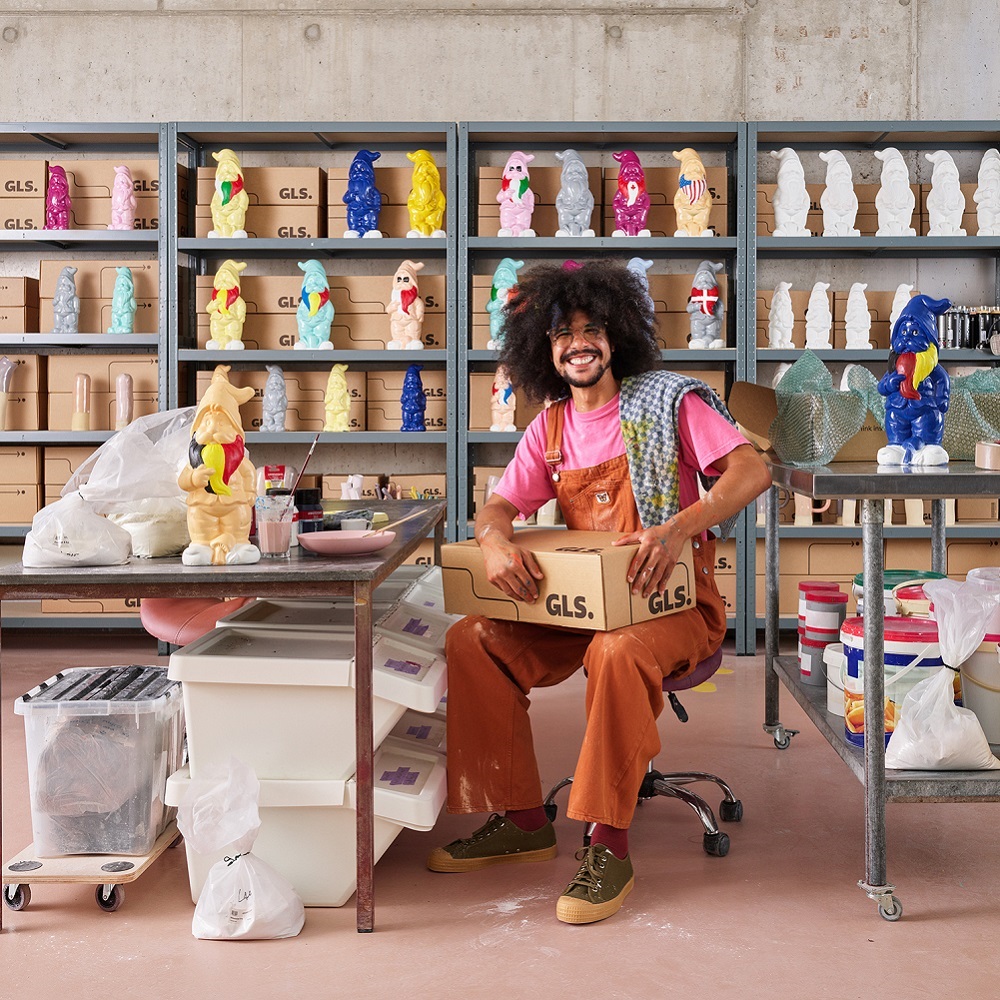 Ein lockiger Mann sitzt mit seinem GLS Paket auf dem Schoß in seinem Atelier