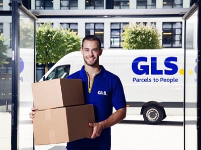 Freundlicher GLS Paketzusteller trägt gerade Pakete durch den Haupteingang in ein Bürkomplex.