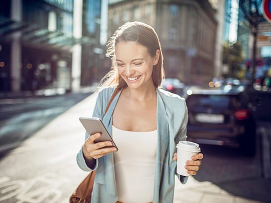 Eine Frau hält einen Coffee to go in der einen Hand und nutzt ihr Smartphone, um mit der anderen Hand die Lieferoption für ihr Paket zu ändern.
